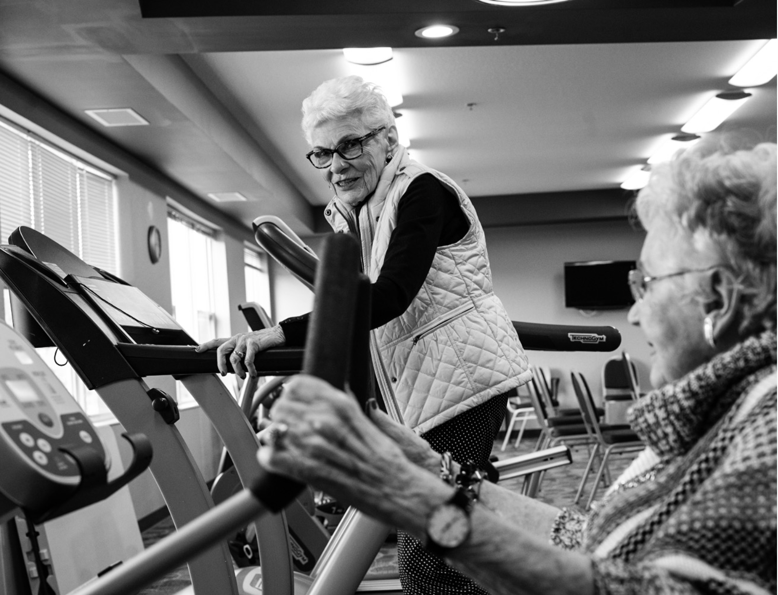 Two female seniors in a fitness room smiling and interacting, one walking on a treadmill, one seated and exercising her arms